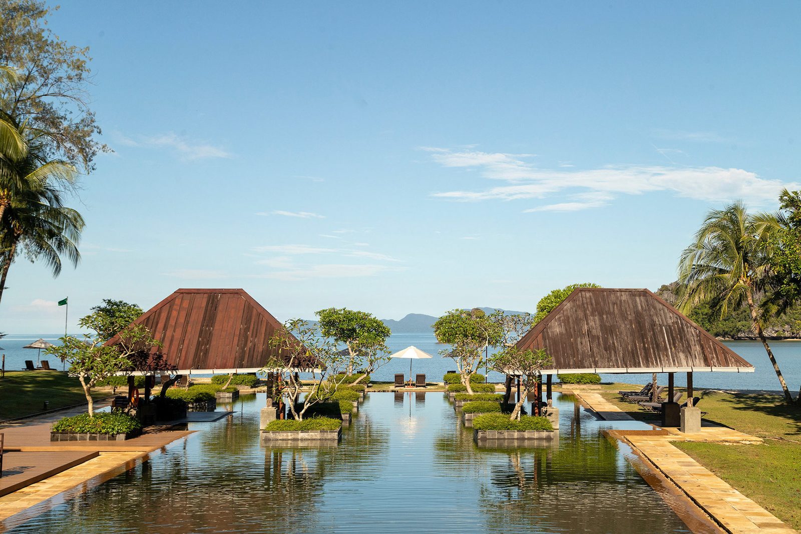Resort water court flanked by twin timber-roofed pavilions, reflecting pool running out to the sea