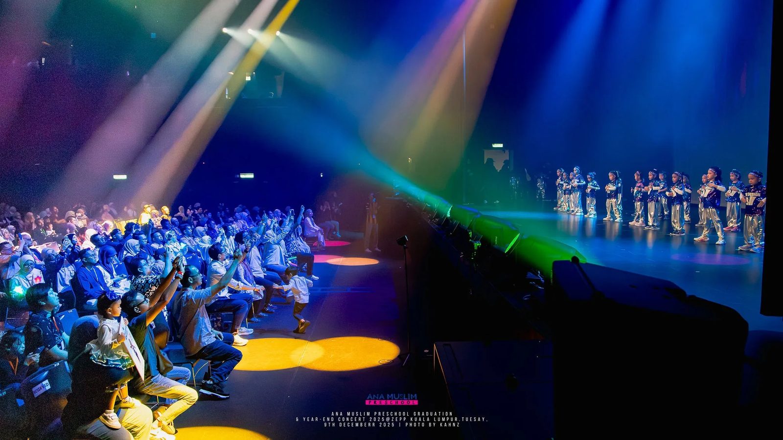 Performers on a large stage under blue and purple beams before a seated crowd