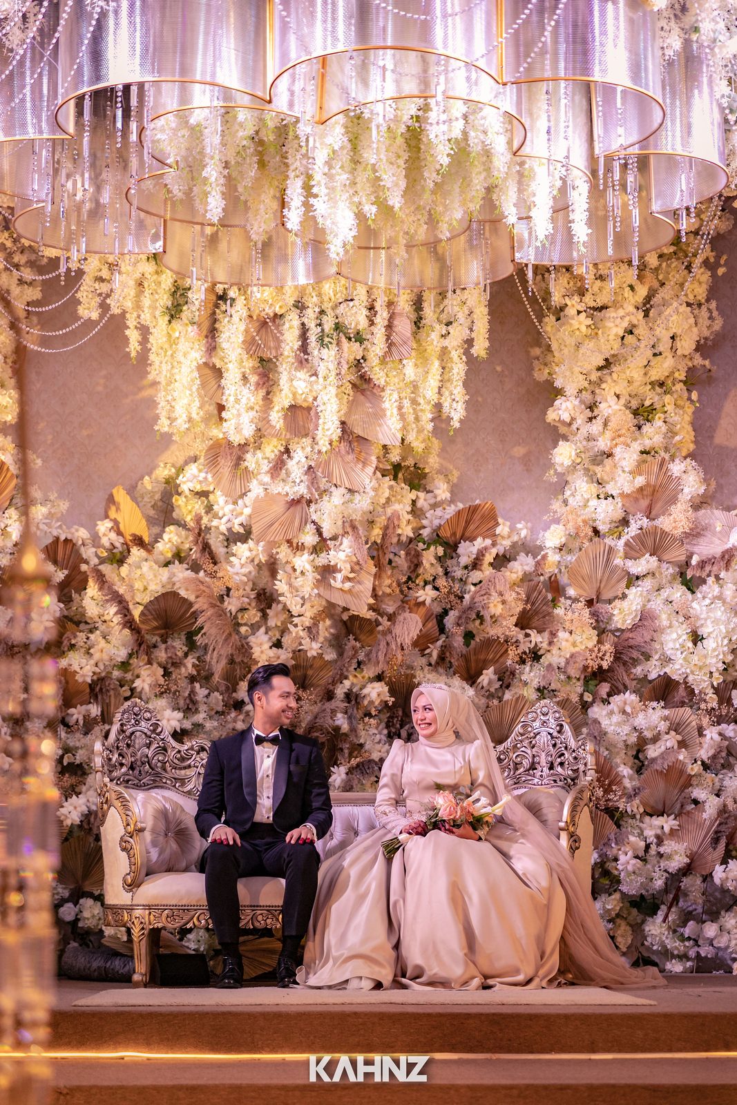Couple seated on an ornate floral dais beneath a chandelier