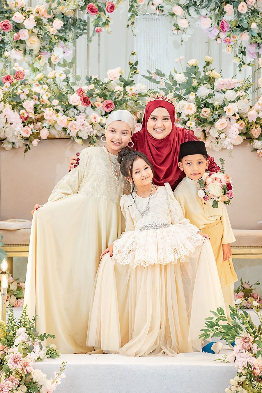 Bride seated with children in cream attire before a floral backdrop